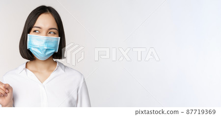 Portrait of cute asian woman in medical face mask, coronavirus protection, pointing finger left and looking intrigued at empty copy space, white background Portrait of cute asian woman in medical face mask, coronavirus protection, pointing finger left and looking intrigued at empty copy space, white background 87719369