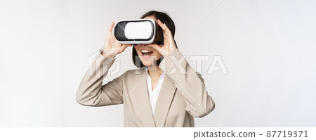 Amazed business woman in suit using virtual reality glasses, looking amazed in vr headset, standing over white background Amazed business woman in suit using virtual reality glasses, looking amazed in vr headset, standing over white background 87719371