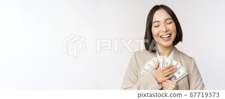 Happy asian businesswoman holding cash, hugging dollars money and smiling, standing over white background in suit Happy asian businesswoman holding cash, hugging dollars money and smiling, standing over white background in suit 87719373