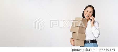 Image of young asian businesswoman answer phone call while carrying boxes for delivery, posing against white background Image of young asian businesswoman answer phone call while carrying boxes for delivery, posing against white background 87719375
