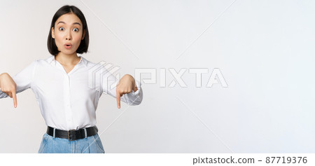 Portrait of young asian woman pointing fingers down and smiling, showing banner, click on link below gesture, inviting people to follow, standing over white background Portrait of young asian woman pointing fingers down and smiling, showing banner, click on link below gesture, inviting people to follow, standing over white background 87719376