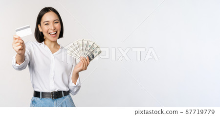 Happy korean woman holding credit card and money dollars, smiling and laughing, posing against white studio background 87719779