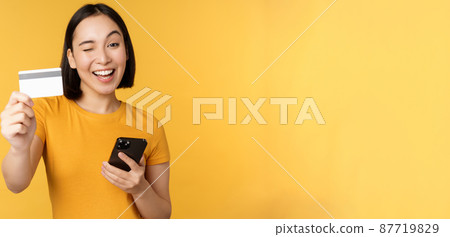 Joyful asian girl smiling, showing credit card and smartphone, recommending mobile phone banking, standing against yellow background 87719829