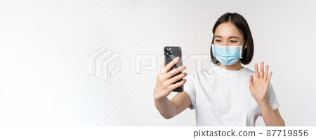 Health and covid-19 concept. Modern asian girl, student in medical mask, video chat with mobile phone, waving hand at smartphone app, standing over white background 87719856