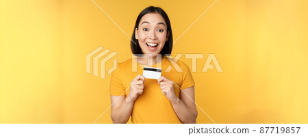 Beautiful asian woman showing credit card and smiling, recommending bank service, standing over yellow background Beautiful asian woman showing credit card and smiling, recommending bank service, standing over yellow background 87719857