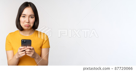 Sad asian woman holding smartphone, looking upset with regret, standing in yellow tshirt against white background 87720066