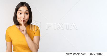Keep quiet. Cute asian woman make shhh gest, showing shush, hush sign, press finger to lips, silence, standing over white background 87720079