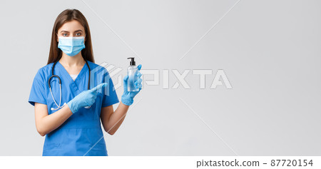 Covid-19, preventing virus, health, healthcare workers and quarantine concept. Young professional female nurse or doctor in blue scrubs, medical mask, pointing finger at hand sanitizer Covid-19, preventing virus, health, healthcare workers and quarantine concept. Young professional female nurse or doctor in blue scrubs, medical mask, pointing finger at hand sanitizer 87720154