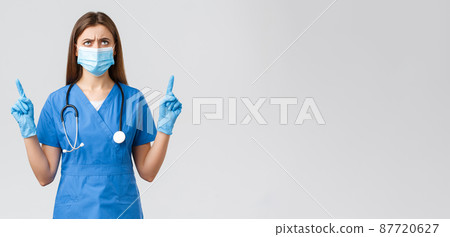 Covid-19, preventing virus, health, healthcare workers and quarantine concept. Concerned and disappointed female nurse or doctor in blue scrubs, medical mask, frowning looking up displeased 87720627