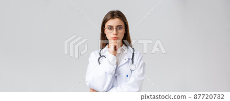 Healthcare workers, medicine, insurance and covid-19 pandemic concept. Serious smart, professional female doctor in glasses and white scrubs, thinking, taking decision or choice, look camera Healthcare workers, medicine, insurance and covid-19 pandemic concept. Serious smart, professional female doctor in glasses and white scrubs, thinking, taking decision or choice, look camera 87720782