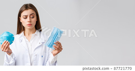 Covid-19, preventing virus, healthcare workers and quarantine concept. Cute female doctor in white scrubs, choosing between medical mask and respirator, pick personal protective equipment Covid-19, preventing virus, healthcare workers and quarantine concept. Cute female doctor in white scrubs, choosing between medical mask and respirator, pick personal protective equipment 87721770