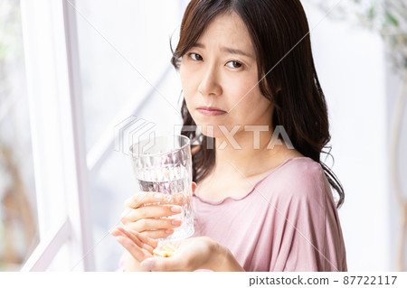 一個穿著休閒服的女人,神情不安,手裡拿著膠囊藥丸和玻璃杯 一個穿著休閒服的女人,神情不安,手裡拿著膠囊藥丸和玻璃杯 87722117