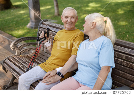 Happy senior man with poles for Nordic walking and wife sit on bench in park Happy senior man with poles for Nordic walking and wife sit on bench in park 87725763