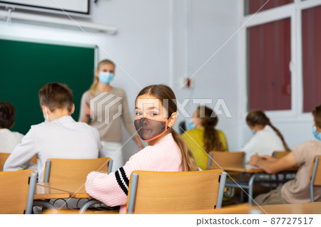 Girl in mask turned around and looking at camera during lesson in classroom Girl in mask turned around and looking at camera during lesson in classroom 87727517
