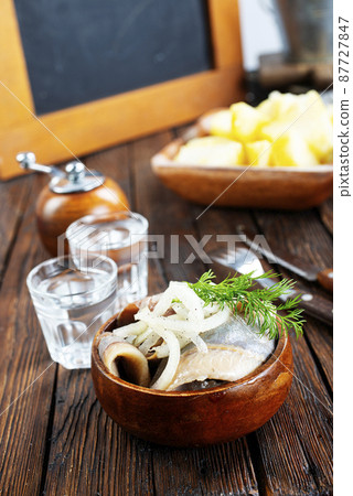 bowl of salted herring on the background of a glass of vodka and decanter. Salted herring in jar as a snack for vodka. bowl of salted herring on the background of a glass of vodka and decanter. Salted herring in jar as a snack for vodka. 87727847