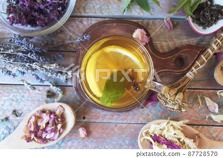 Cup of green tea with mint and lemon, dry petals of useful plants on a wooden table Cup of green tea with mint and lemon, dry petals of useful plants on a wooden table 87728570