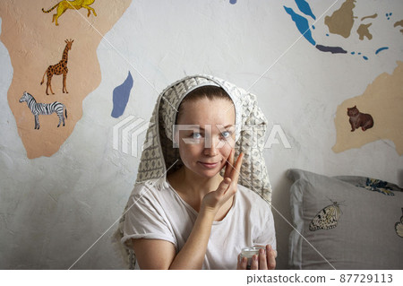 An attractive happy woman of thirty years smears her cheeks with cream, a towel on her head, a home beauty ritual every day 87729113