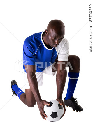 African american young male soccer player kneeling by ball against white background African american young male soccer player kneeling by ball against white background 87730780