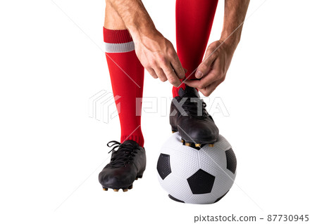 Low section of young male caucasian player tying shoelace on soccer ball over white background 87730945