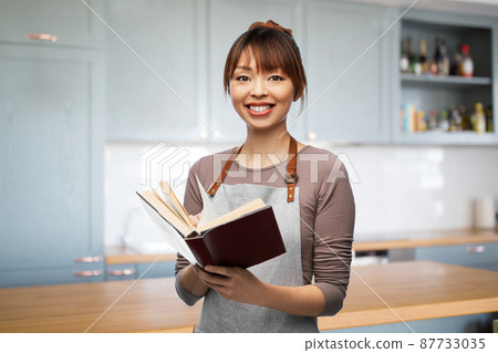 smiling woman in apron with cook book at kitchen 87733035