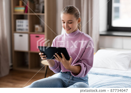 smiling girl with tablet pc sitting on bed at home smiling girl with tablet pc sitting on bed at home 87734047