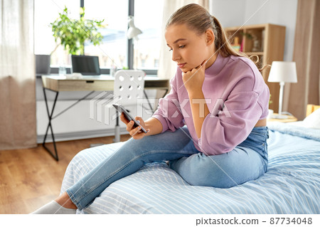 sad girl with smartphone sitting on bed at home sad girl with smartphone sitting on bed at home 87734048