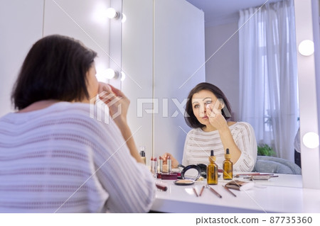 Beautiful middle aged woman doing makeup in front of a mirror. Beautiful middle aged woman doing makeup in front of a mirror. 87735360