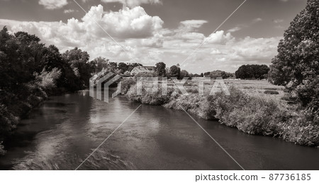 Landscape with a river next to a field and a farm with clouds in the background, black and white Landscape with a river next to a field and a farm with clouds in the background, black and white 87736185