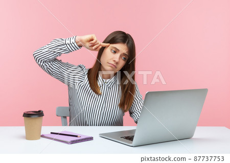 Depressed unhappy woman office worker in striped shirt sitting workplace showing stupid gesture to laptop display talking on video call. Indoor studio shot isolated on pink background Depressed unhappy woman office worker in striped shirt sitting workplace showing stupid gesture to laptop display talking on video call. Indoor studio shot isolated on pink background 87737753
