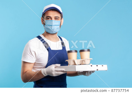 Delivery service. Friendly professional courier in uniform and mask holding coffee and pizza box, wearing safety gloves offering drinks food, looking at camera with toothy smile. studio shot isolated Delivery service. Friendly professional courier in uniform and mask holding coffee and pizza box, wearing safety gloves offering drinks food, looking at camera with toothy smile. studio shot isolated 87737754