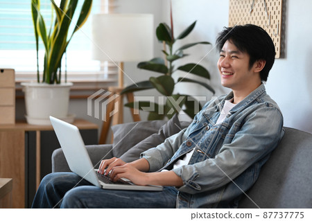 Cheerful young asian man using laptop computer while sitting on couch at home. Cheerful young asian man using laptop computer while sitting on couch at home. 87737775