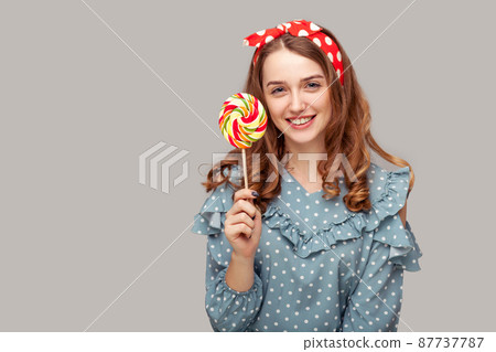 Pretty pinup girl ruffle blouse holding sweet spiral candy looking at camera, smiling with delicious confectionery lollipop in her hands. retro vintage style. studio shot isolated gray background, Pretty pinup girl ruffle blouse holding sweet spiral candy looking at camera, smiling with delicious confectionery lollipop in her hands. retro vintage style. studio shot isolated gray background, 87737787