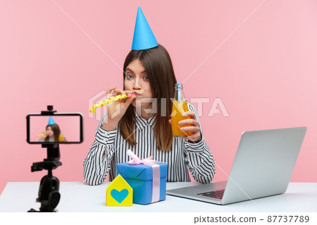 Funny woman in cone hat blowing at party blower holding bottle with lemonade, recording video congratulation on smartphone camera, preparing gifts. Indoor studio shot isolated on pink background Funny woman in cone hat blowing at party blower holding bottle with lemonade, recording video congratulation on smartphone camera, preparing gifts. Indoor studio shot isolated on pink background 87737789