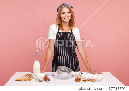Happy smiling female preparing bakery, making cookies, preparing ingredients for baking pasty, looking at camera, wearing striped apron. Indoor studio shot isolated on pink background. Happy smiling female preparing bakery, making cookies, preparing ingredients for baking pasty, looking at camera, wearing striped apron. Indoor studio shot isolated on pink background. 87737791