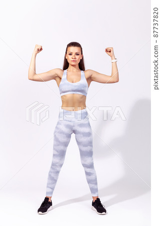 Full length self confident athletic woman showing her strong abs and biceps raising hands up, doing sport, pumping up muscles. Indoor studio shot isolated on gray background 87737820