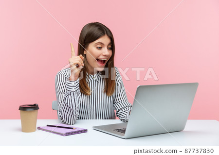 Smiling positive woman office worker in striped shirt pointing finger up talking video call on laptop, great idea for startup, business solution. Indoor studio shot isolated on pink background 87737830