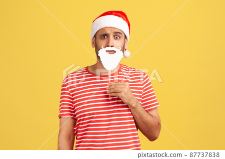 Thoughtful confused man in red t-shirt holding paper beard near chin and wearing santa claus hat, puzzled with fancy-dress party. Indoor studio shot isolated on yellow background 87737838