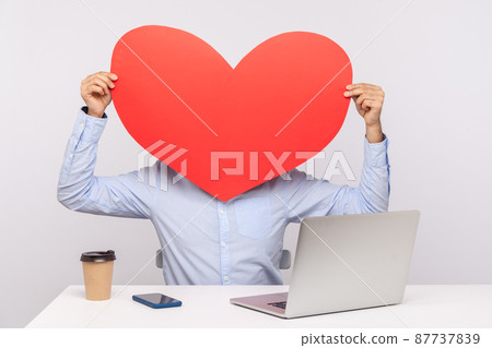 Man in shirt sitting at desk laptop, hiding face behind red heart, expressing affection, romantic feelings, holding huge greeting card on Valentine's Day. studio shot isolated on white background 87737839