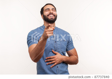 Extremely happy bearded man laughing holding hand on belly and pointing finger at you looking at camera, funny ridiculous joke. Indoor studio shot isolated on white background 87737840