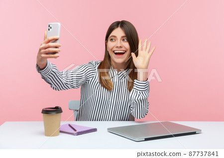 Smiling positive woman in striped shirt saying hello and waving hand looking at smartphone camera, keeping online video call, recording video. Indoor studio shot isolated on pink background 87737841