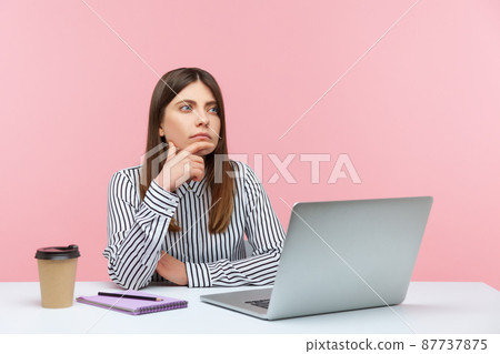 Pensive brunette woman in striped shirt sitting with thoughtful confused expression while working on laptop at home office, thinking over business plan. Indoor studio shot isolated on pink background Pensive brunette woman in striped shirt sitting with thoughtful confused expression while working on laptop at home office, thinking over business plan. Indoor studio shot isolated on pink background 87737875