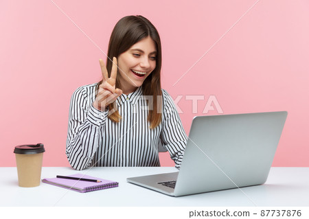 Successful positive woman office worker showing victory gesture and smiling looking into laptop screen, talking video call, online conference. Indoor studio shot isolated on pink background Successful positive woman office worker showing victory gesture and smiling looking into laptop screen, talking video call, online conference. Indoor studio shot isolated on pink background 87737876