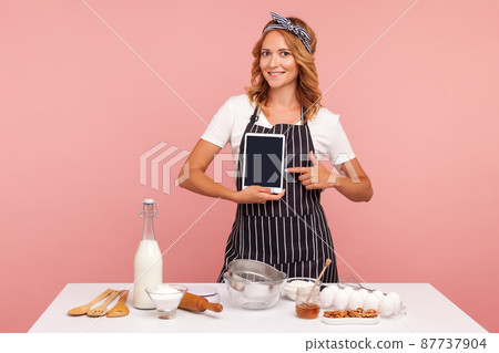 Portrait of beautiful young adult woman holding tablet computer and pointing at it blank screen while standing near table with bakery ingredients. Indoor studio shot isolated on pink background. 87737904