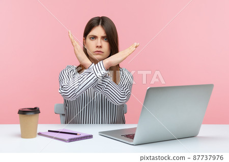 Upset bossy woman office worker sitting at workplace crossing hands showing x sign, rejecting and finishing sexual harassment at work. Indoor studio shot isolated on pink background Upset bossy woman office worker sitting at workplace crossing hands showing x sign, rejecting and finishing sexual harassment at work. Indoor studio shot isolated on pink background 87737976