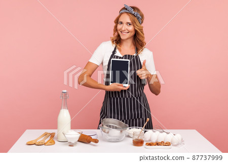 Portrait of attractive young adult female in apron cooking bakery, holding blank tablet in hand and showing thumb up, looking at camera with smile. Indoor studio shot isolated on pink background. 87737999