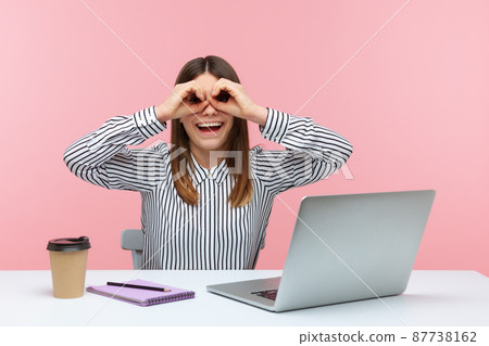 Smiling positive woman office worker sitting at workplace holding hands near eyes pretending to look in binoculars, trying to find someone. Indoor studio shot isolated on pink background Smiling positive woman office worker sitting at workplace holding hands near eyes pretending to look in binoculars, trying to find someone. Indoor studio shot isolated on pink background 87738162