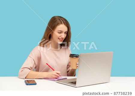 Woman drinking coffee, working on laptop and making notes on paper. Student studying, writing in notebook while listening online school, internet course. indoor studio shot isolated on blue background 87738239