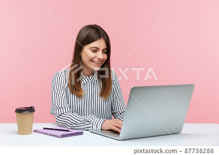 Positive woman office worker in striped shirt sitting on workplace with smile, drinking coffee and making notes working on laptop, good mood. Indoor studio shot isolated on pink background 87738288