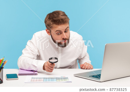 Nosy man office worker with beard looking at accounts data through magnifying glass, spying, finding out information sitting at workplace with laptop. Indoor studio shot isolated on blue background Nosy man office worker with beard looking at accounts data through magnifying glass, spying, finding out information sitting at workplace with laptop. Indoor studio shot isolated on blue background 87738325