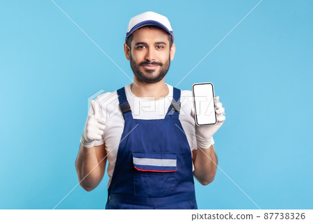 Satisfied consultant in overalls and gloves holding mobile phone with mock up display and showing thumbs up. Good online delivery order app, house repair maintenance services. indoor studio shot Satisfied consultant in overalls and gloves holding mobile phone with mock up display and showing thumbs up. Good online delivery order app, house repair maintenance services. indoor studio shot 87738326